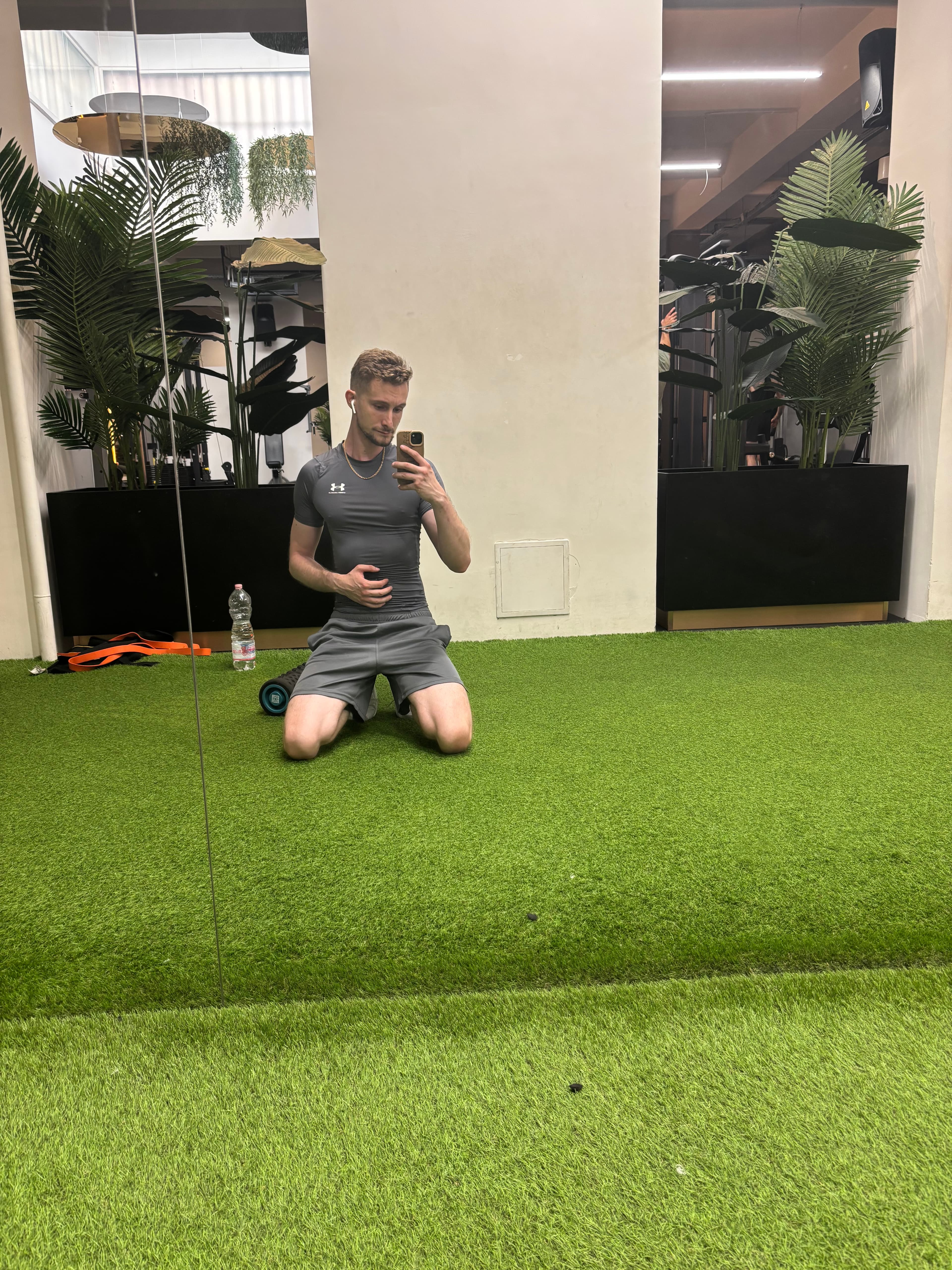 Man in grey athletic wear taking a mirror selfie while kneeling on green artificial turf.